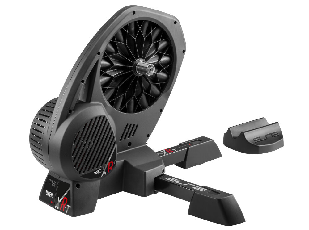 Na obrazie znajduje się czarny trenażer rowerowy z widocznymi napisami \'DIRETO XR-T\' i \'ELITE\'. Obok trenażera leży podkładka z napisem \'ELITE\'.