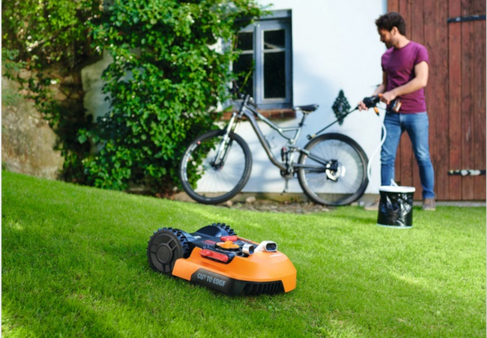 Robot koszący WORX Landroid S250 trawnik robot na trawie w tle meżczyzna rower dom automatyczne koszenie zadbany trawnik koszenie bez wysiłku zdalne zarządzanie
