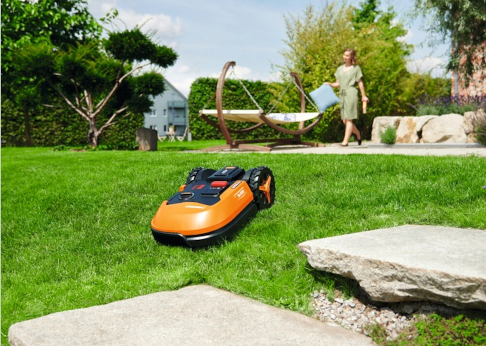 Robot koszący WORX Landroid S250 trawnik robot jedzie po wzniesieniu w tle kobieta dom koszenie na wzniesianiach o nachyleniu terenu do 19 stopni nie traci przyczepności zachowuje stabilność