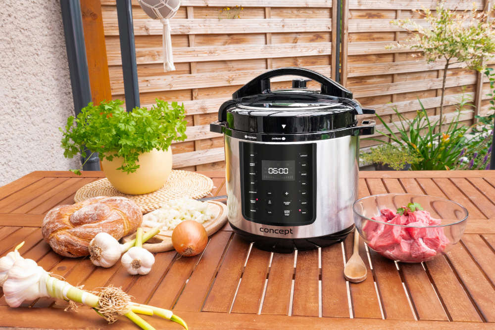Na drewnianym stole znajduje się multicooker z wyświetlaczem pokazującym \'05:00\', obok którego leżą składniki takie jak chleb, czosnek, cebula i miska z mięsem. W tle widoczna jest drewniana kratka ogrodowa oraz rośliny.