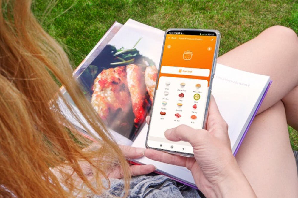 Osoba trzyma smartfon z aplikacją do gotowania, na ekranie widoczne są ikony różnych potraw. W tle otwarta książka kucharska z fotografią dania.