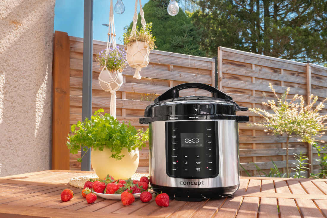 Na drewnianym stole stoi multicooker z wyświetlaczem pokazującym \'06:00\', obok którego leży kilka truskawek i doniczka z ziołami. W tle widoczny jest drewniany płot i wiszące doniczki z roślinami.