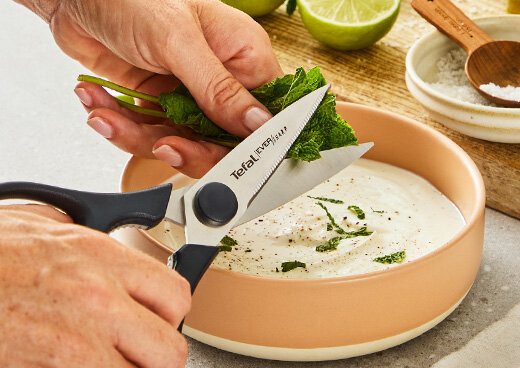 Zbliżenie na dłoń używającą nożyc kuchennych Tefal EverSharp K2977S44 do krojenia świeżej mięty nad miską z jogurtem.