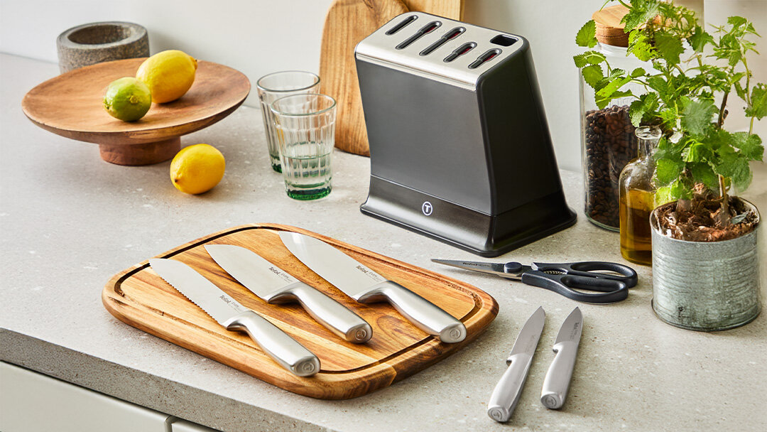 Zestaw noży K2977S44 rozłożony na desce do krojenia i na blacie. Obok blok z automatyczną ostrzałką i inne akcesoria.
