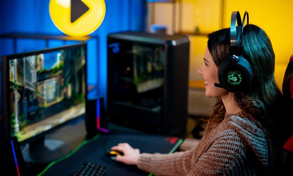 Komputer MAD DOG MD-I02DR32 Kobieta w słuchawkach siedząca przy biurku z komputerem i monitorem w pokoju oświetlonym niebieskim i żółtym światłem