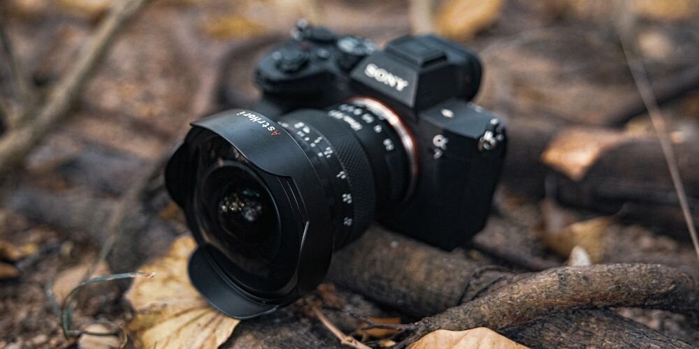 Obiektyw fisheye widziany z boku z widocznymi pierścieniami regulacji ostrości i przysłony oraz oznaczeniami na tle miasta nocą i wzgórz o zachodzie słońca oraz logo AstrHori