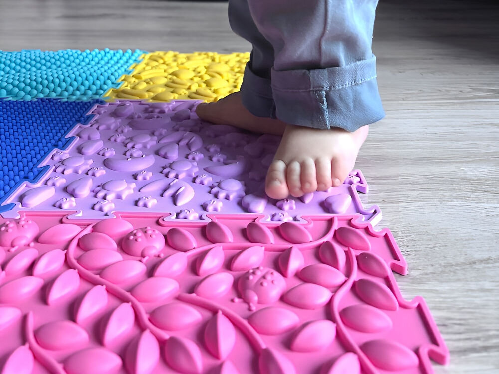 Mata sensoryczna ORTOPEDEK 128924 24.5 x 24.5 cm (6 elementów) Wielokolorowy Zbliżenie na błękitny, kwadratowy element maty sensorycznej Ortopedek o strukturze drobnych, masujących igiełek. W centralnej części widoczne wypukłe logo marki Ortopedek.