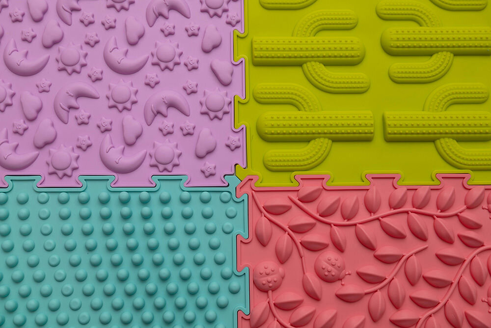 Mata sensoryczna ORTOPEDEK 128924 24.5 x 24.5 cm (6 elementów) Wielokolorowy Makrofotografia pastelowego, różowego puzzla ortopedycznego Ortopedek. Zbliżenie pokazuje detale wypukłego wzoru w kształcie stóp z logotypem marki, służącego do stymulacji receptorów dotykowych.