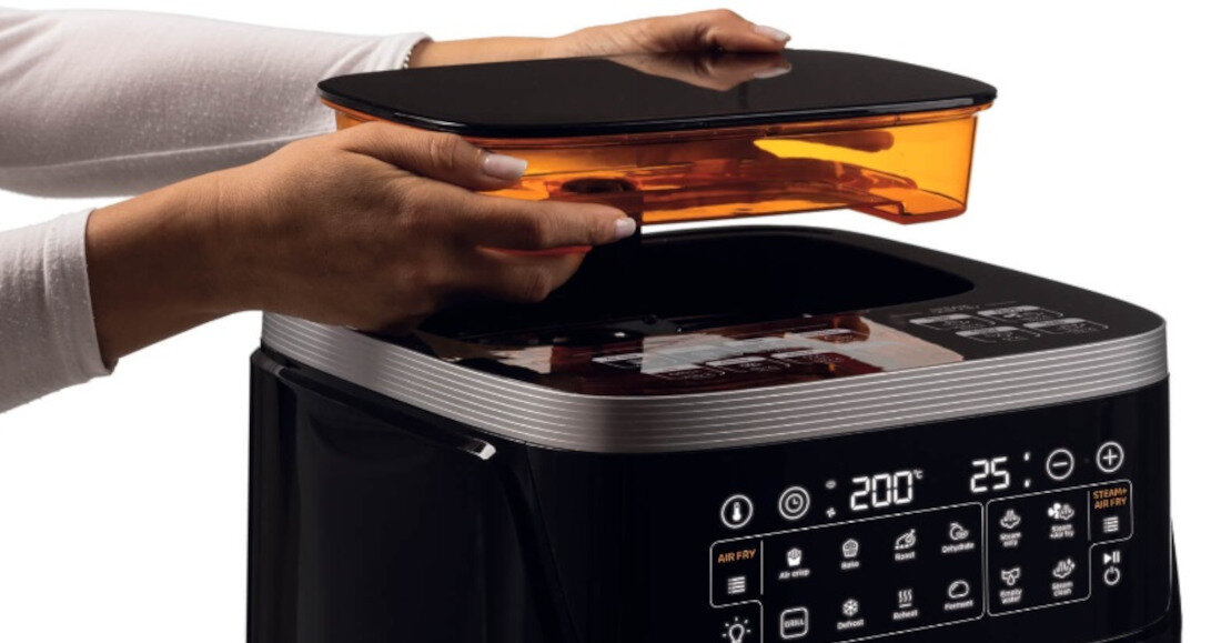 Osoba trzyma pomarańczową pokrywę nad czarną frytkownicą beztłuszczową z wyświetlaczem cyfrowym pokazującym temperaturę 200°C i czas 25 minut. Na panelu sterowania widoczne są różne ikony funkcji gotowania.