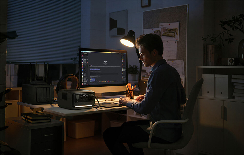 Stacja zasilania ECOFLOW River 3 Plus 286 Wh pokój osoba sidzie przy biurku na biurku stacja zasilania laptop i lampka trzy gniazda AC o mocy wyjściowej 600 W możliwość zwiększenia do 1200 W dzięki trybowi X-Boost dwa porty USB-A o łącznej mocy 24 W port USB-C o mocy 100 W gniazdo samochodowe 12,6 V o mocy 126 W