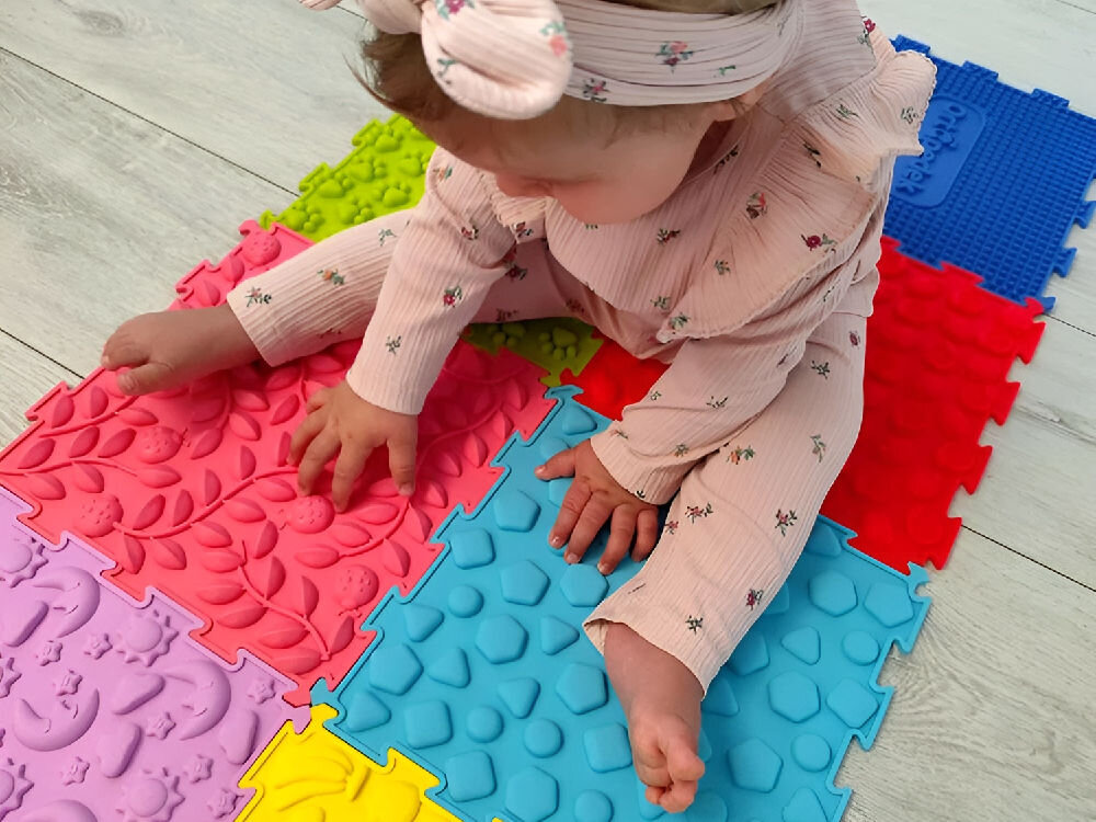 Mata sensoryczna ORTOPEDEK 128931 24.5 x 24.5 cm (8 elementów) WielokolorowyMata sensoryczna ORTOPEDEK 128931 24.5 x 24.5 cm (8 elementów) Wielokolorowy Dziecko bawiące się na podłodze pastelowymi puzzlami sensorycznymi Ortopedek. Widoczne dłonie dotykające niebieskiego elementu z drobnymi wypustkami i logo marki oraz różowego puzzla ze wzorem stóp.