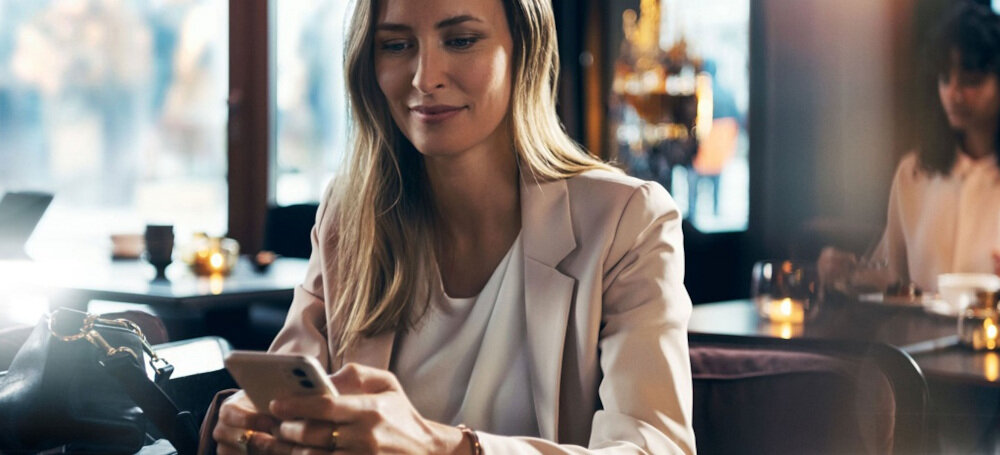 ZMYWARKA SIEMENS SN63EX03ME Zdjęcie ukazuje kobietę korzystającą ze smartfona w eleganckim wnętrzu, co symbolizuje możliwość zdalnego sterowania zmywarką. Ujęcie jest jasne, stylizowane i podkreśla wygodę użytkowania Home Connect sterowanie zdalne aplikacja smartfon inteligentny dom zdalna diagnostyka optymalizacja energii