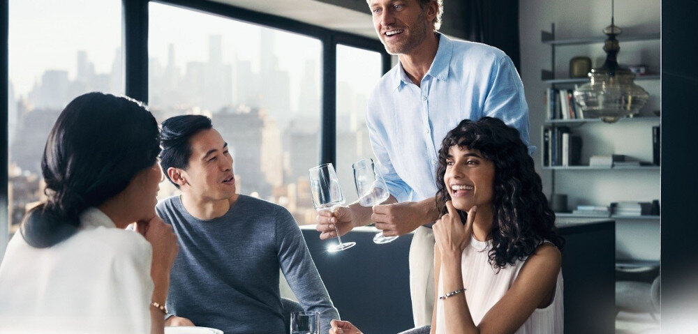 ZMYWARKA SIEMENS SN63EX03ME Zdjęcie przedstawia grupę czterech osób siedzących przy stole w jasnym wnętrzu z dużymi oknami i widokiem na miasto. Atmosfera jest swobodna i radosna — jedna z osób trzyma kieliszki, jakby wznoszono toast lub rozpoczynano spotkanie towarzyskie glassCare ochrona szkła delikatne mycie kieliszki pielęgnacja szkła połysk naczyń