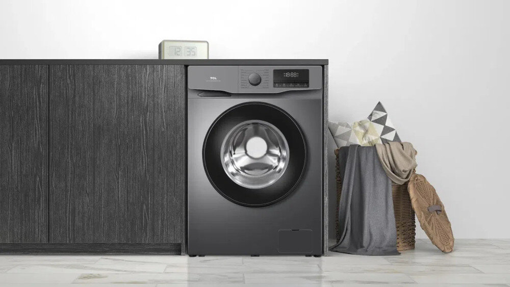 PRALKA TCL FF0612SB1PL Na pierwszym zdjęciu biała pralka ładowana od frontu została wbudowana w ciemną, grafitową zabudowę z imitacją drewna, tworząc elegancki, minimalistyczny wystrój. Obok stoją kosze z poduszkami i kocem, które ocieplają aranżację domowej pralni.