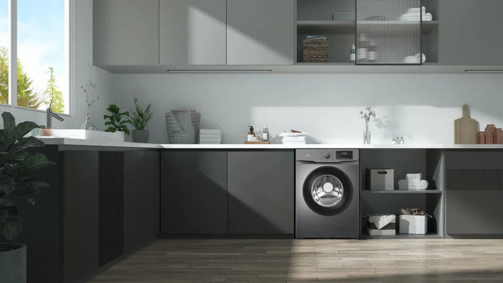 PRALKA TCL FF0612SB1PL Na drugim zdjęciu pralka jest centralnie umieszczona w nowoczesnej zabudowie z szarymi frontami i czarnym blatem, pełniąc rolę głównego elementu strefy pralni. Nad blatem znajduje się podświetlana szafka z półkami na kosmetyki i detergenty oraz dekoracje, a po prawej stronie widoczny jest regał i duża roślina doniczkowa na drewnianej podłodze energooszczędność cicha praca inteligentne technologie komfort