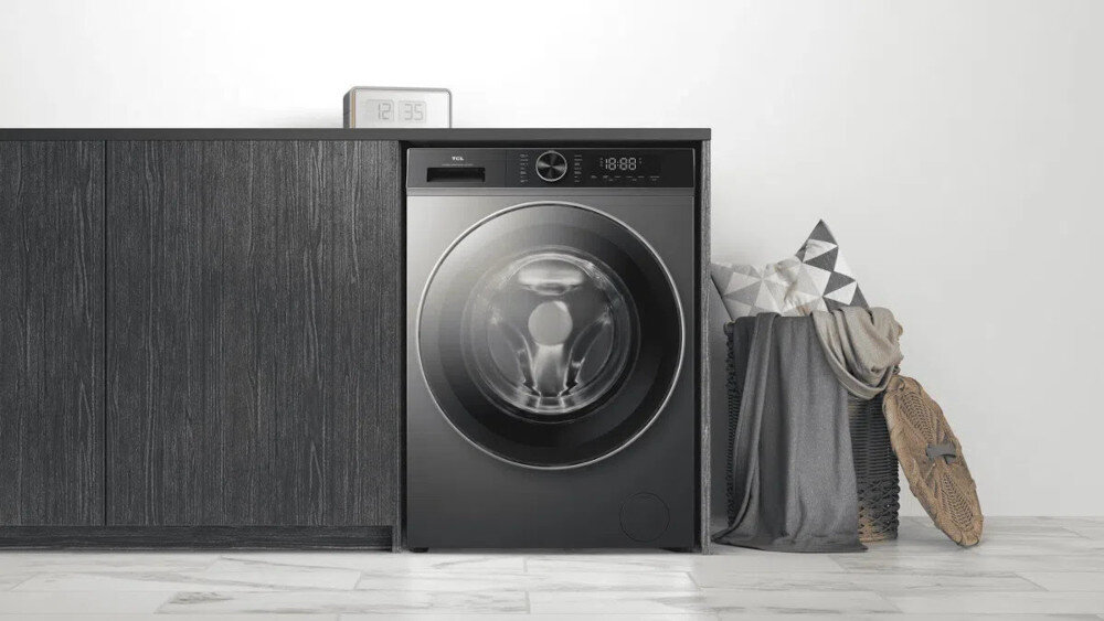 PRALKA TCL FP0834SA1PL Ciemna pralka zabudowana w ciągu szafek z ciemnego drewna, z zegarem ustawionym na blacie oraz koszem z poduszkami i kocem po prawej stronie, stojąca na jasnej podłodze