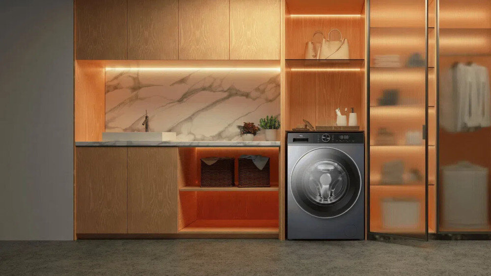 PRALKA TCL FP0834SA1PL Ciemna pralka wolnostojąca wkomponowana w zabudowę z czarnym blatem, otoczona wiszącą szafką z podświetlanymi półkami, rośliną w donicy, regałem oraz koszem na pranie, tworząca przytulną aranżację pralni energooszczędna inteligentne funkcje higieniczne pranie Steam Wash Auto Weight Honeycomb Drum czyste ubrania świeżość bezpieczeństwo komfort jakość