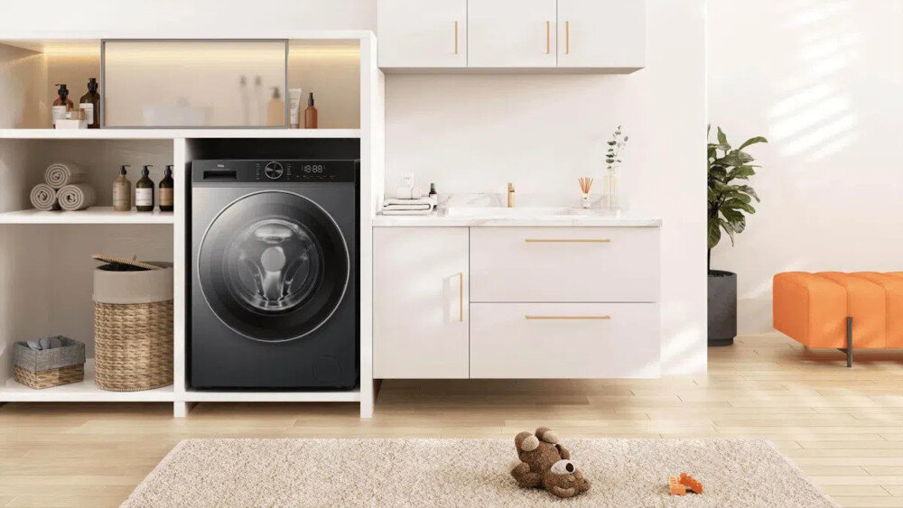 PRALKA TCL FP0834SA1PL Ciemna pralka wstawiona w białą zabudowę z otwartymi półkami na ręczniki i kosmetyki, obok szafki z umywalką, rośliną oraz pomarańczowym siedziskiem, na dywanie leży pluszowy miś