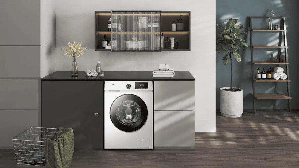 PRALKA TCL FF0824WA1PL Pralka zabudowana w eleganckiej zabudowie z ciemnym blatem, otoczona szafkami, otwartymi półkami z kosmetykami oraz rośliną i regałem z ręcznikami po prawej stronie