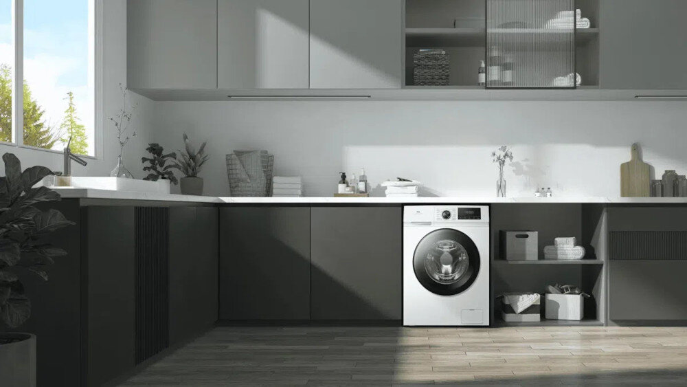 PRALKA TCL FF0824WA1PL Pralka wkomponowana w jasną, przestronną zabudowę kuchenną w kształcie litery L, z dużym oknem, licznymi szafkami, dekoracyjnymi roślinami i akcesoriami do prania na blacie oraz półkach cicha praca higiena prania wygoda użytkowania łazienka