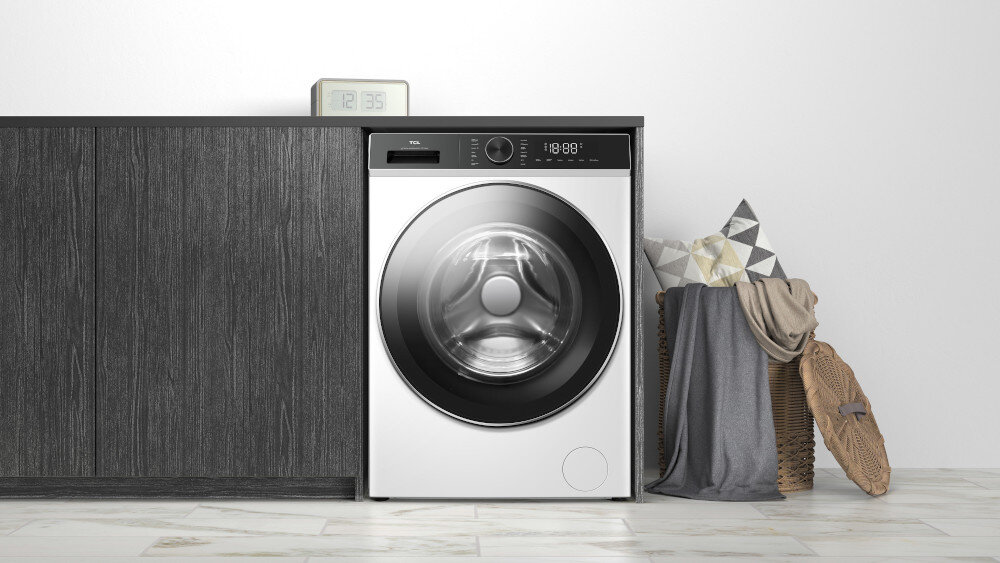 PRALKA TCL FP0834WA1PL Biała pralka zabudowana w ciągu szafek z ciemnego drewna, z zegarem ustawionym na blacie oraz koszem z poduszkami i kocem po prawej stronie, stojąca na jasnej podłodze