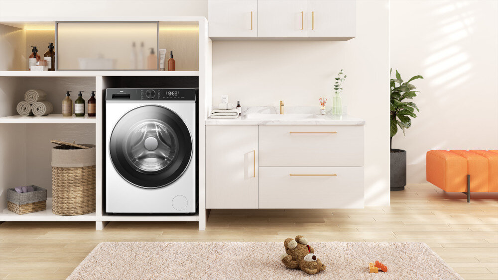 PRALKA TCL FP0834WA1PL Biała pralka wstawiona w białą zabudowę z otwartymi półkami na ręczniki i kosmetyki, obok szafki z umywalką, rośliną oraz pomarańczowym siedziskiem, na dywanie leży pluszowy miś