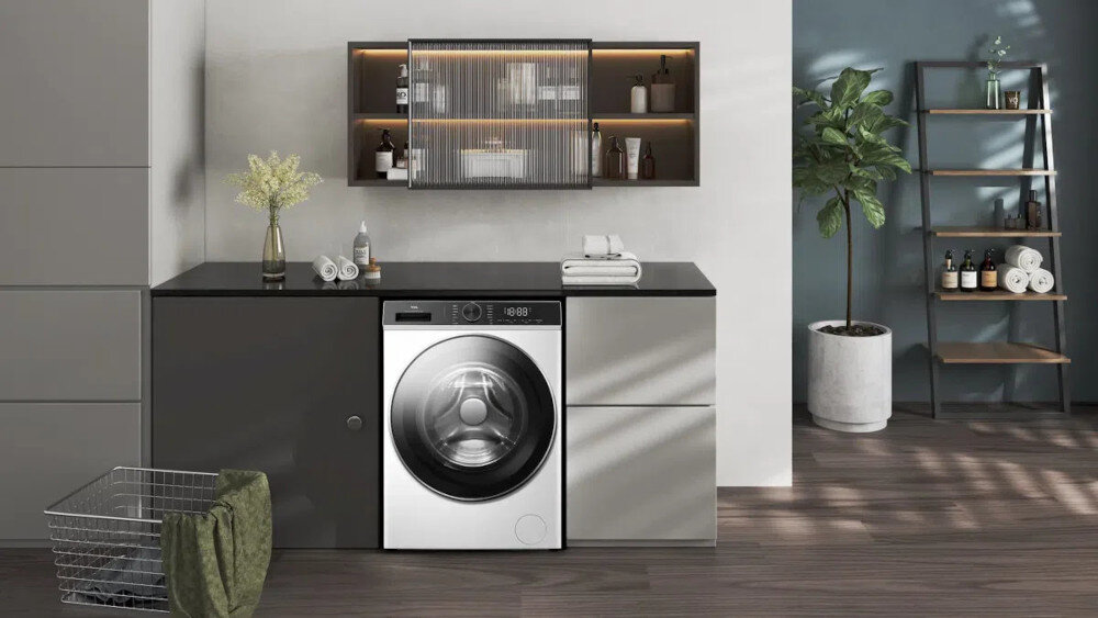 PRALKA TCL FP0934WA1PL Na drugim zdjęciu pralka jest centralnie umieszczona w nowoczesnej zabudowie z szarymi frontami i czarnym blatem, pełniąc rolę głównego elementu strefy pralni. Nad blatem znajduje się podświetlana szafka z półkami na kosmetyki i detergenty oraz dekoracje, a po prawej stronie widoczny jest regał i duża roślina doniczkowa na drewnianej podłodze energooszczędność cicha praca inteligentne technologie komfort