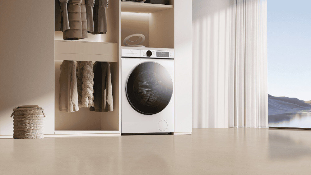 PRALKA TCL FP0944WA0PL Minimalistyczne wnętrze z zabudowaną szafą pokazuje białą pralko‑suszarkę ustawioną we wnęce obok wieszaka z ubraniami, z dużym przeszklonym bębnem na froncie i widokiem na jasną, słoneczną przestrzeń za zasłoną