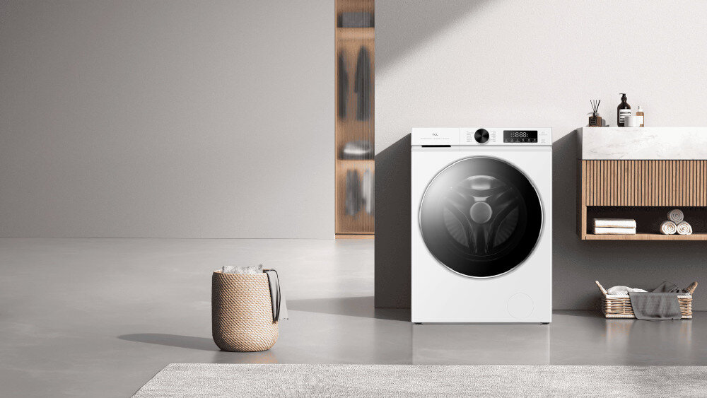 PRALKA TCL FP0944WA0PL Biała pralko‑suszarka stoi na tle eleganckiej łazienki z wiszącą szafką pod umywalką i drewnianymi lamelami, obok kosza na pranie i wiklinowego koszyka z ręcznikami, co podkreśla nowoczesny, spa‑like charakter pomieszczenia inteligentne technologie energooszczędność Steam Wash MAGIC Flow eliminacja bakterii system antybakteryjny higiena świeżość ubrań bezpieczeństwo skóry