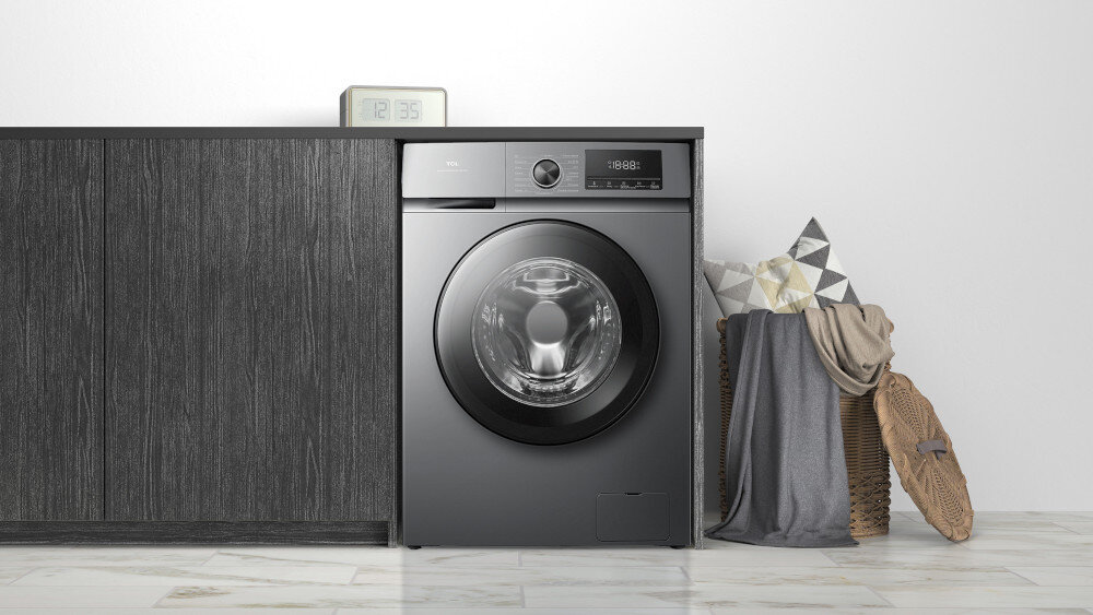 PRALKA TCL FF0924SA0PL Pralka w ciemnym, grafitowym kolorze wkomponowana w zabudowę z czarnymi frontami, obok kosz z poduszkami i kocem oraz zegar na blacie, tworzące spójną aranżację pralni