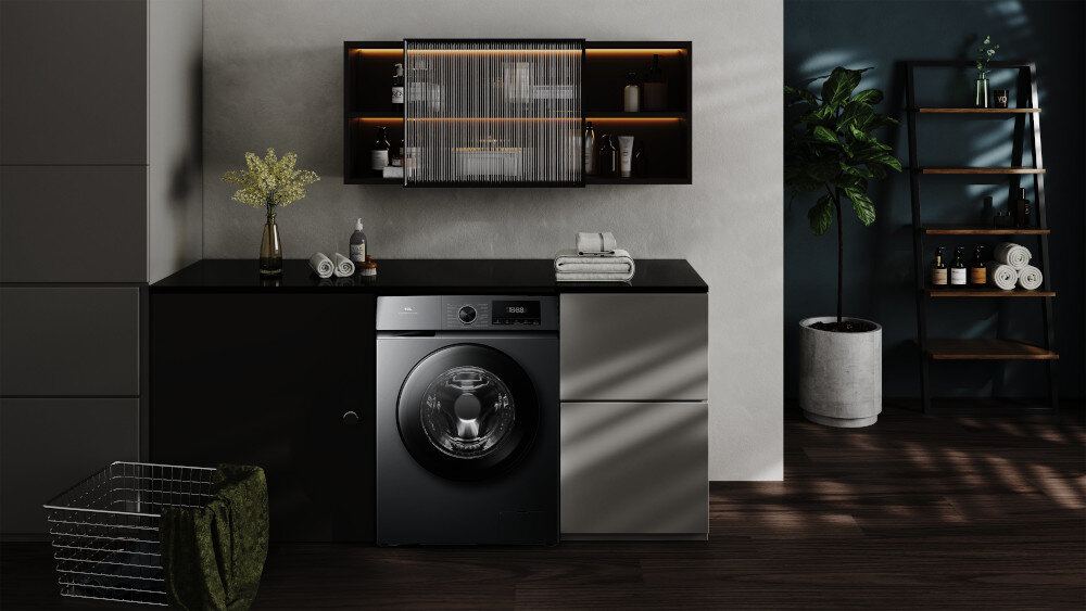 PRALKA TCL FF0924SA0PL Ciemna pralka wolnostojąca wkomponowana w zabudowę z czarnym blatem, otoczona wiszącą szafką z podświetlanymi półkami, rośliną w donicy, regałem oraz koszem na pranie, tworząca przytulną aranżację pralni energooszczędna inteligentne funkcje higieniczne pranie Steam Wash Auto Weight Honeycomb Drum czyste ubrania świeżość bezpieczeństwo komfort jakość