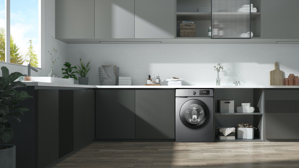 PRALKA TCL FF0924SA0PL Pralka w ciemnym kolorze wbudowana w zabudowę w kształcie litery L, otoczona szarymi szafkami, roślinami i akcesoriami do prania, w jasnym, przestronnym pomieszczeniu z dużym oknem energooszczędność cicha praca higiena prania wygoda użytkowania łazienka