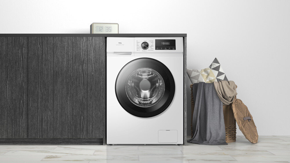 PRALKA TCL FF0924WA0PL Na pierwszym zdjęciu biała pralka ładowana od frontu została wbudowana w ciemną, grafitową zabudowę z imitacją drewna, tworząc elegancki, minimalistyczny wystrój. Obok stoją kosze z poduszkami i kocem, które ocieplają aranżację domowej pralni.