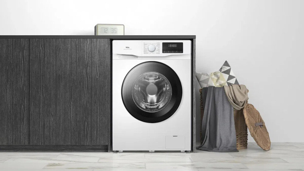 PRALKA TCL FF0714WA1PL Pralka ładowana od frontu stoi we wnęce między ciemnymi, grafitowymi szafkami a koszem z poduszkami i kocami po prawej stronie. Na blacie nad urządzeniem ustawiono prosty zegar elektroniczny, a całość otacza jasna, jednolita ściana i jasna podłoga z dużych płytek