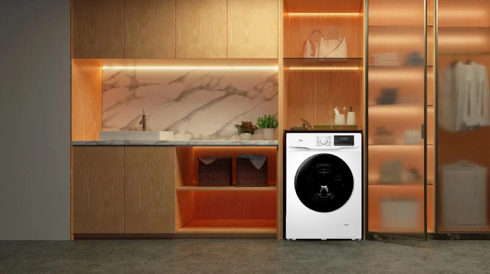 PRALKA TCL FF0714WA1PL 2.jpg: Na zdjęciu widoczna jest biała pralka ładowana od frontu ustawiona w zabudowie z jasnego drewna, obok otwartych półek z koszami i dekoracjami. Tło stanowią podświetlane wnęki z ciepłym światłem oraz fragment przeszklonej szafy, co podkreśla elegancki charakter domowej pralni cicha praca innowacyjne technologie higiena prania łazienka pojemność 7kg