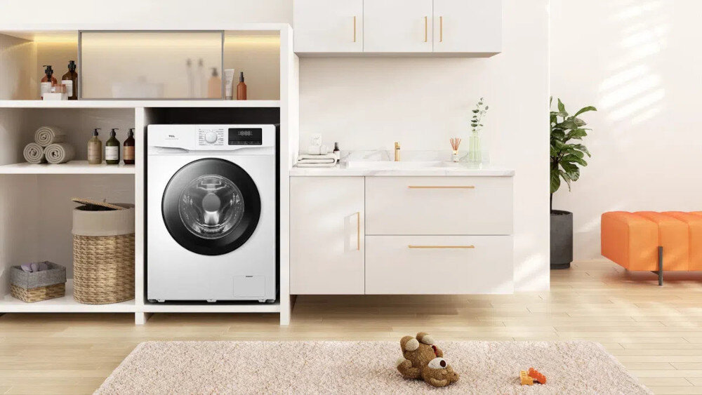 PRALKA TCL FF0714WA1PL Pralka ładowana od frontu jest zabudowana w jasnej łazience z białymi szafkami i otwartymi półkami, na których stoją ręczniki, kosmetyki oraz kosz z wikliny. Na pierwszym planie leży beżowy dywanik z pluszowym misiem, a w głębi widać pomarańczową ławkę i roślinę w donicy cicha praca spokój