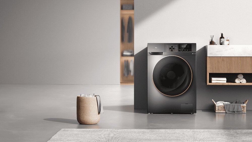 PRALKO-SUSZARKA TCL CP1012SA0PL Ciemna pralka z dużymi, przyciemnianymi drzwiami w miedzianej ramce stoi obok umywalki z szafką o lamelowym froncie, na tle jasnej ściany; obok znajdują się kosz na pranie i akcesoria łazienkowe