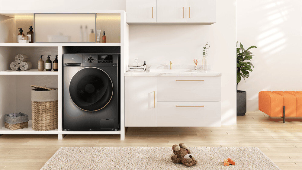 PRALKO-SUSZARKA TCL CP1012SA0PL Ciemna pralka z dużymi drzwiami ozdobionymi miedzianą ramką, wkomponowana w białą zabudowę z otwartymi półkami na ręczniki i kosmetyki, obok szafki z umywalką, rośliną i pomarańczowym siedziskiem oraz pluszowym misiem na dywanie pralko-suszarka pranie suszenie kompaktowa forma Steam Wash Wash & Dry 60' dodawanie ubrań wygoda oszczędność miejsca