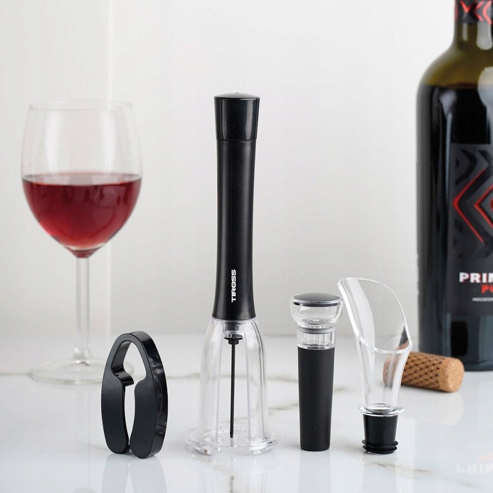 Na obrazie znajduje się zestaw akcesoriów do wina, w tym korkociąg ciśnieniowy, obcinacz do folii, nalewak i korek. Obok zestawu stoi kieliszek z czerwonym winem oraz butelka wina z widocznym korkiem na blacie.