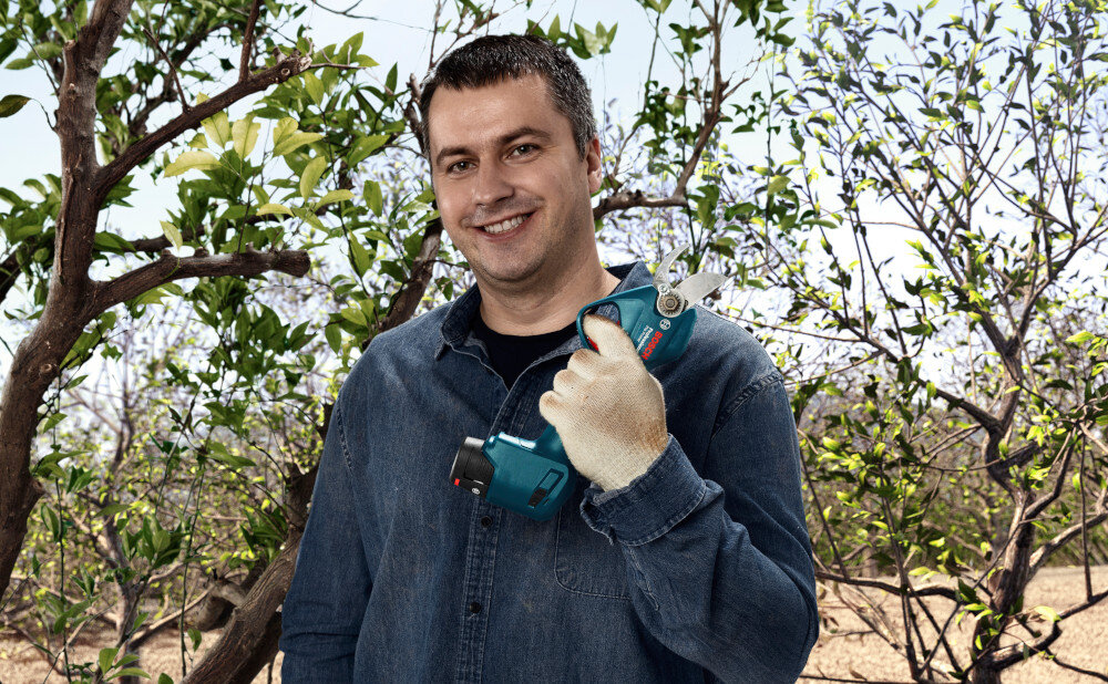 Sekator akumulatorowy BOSCH Pro Pruner 06019K1021 ogród drzewa sad niebo uśmiechnięta osoba trzyma akumulator w dłoni waga 0,8 kg bez akumulatora waga 1 kg z zamontowaną baterią długość całkowita 302 mm ergonomiczna konstrukcja dobrze leży w dłoni