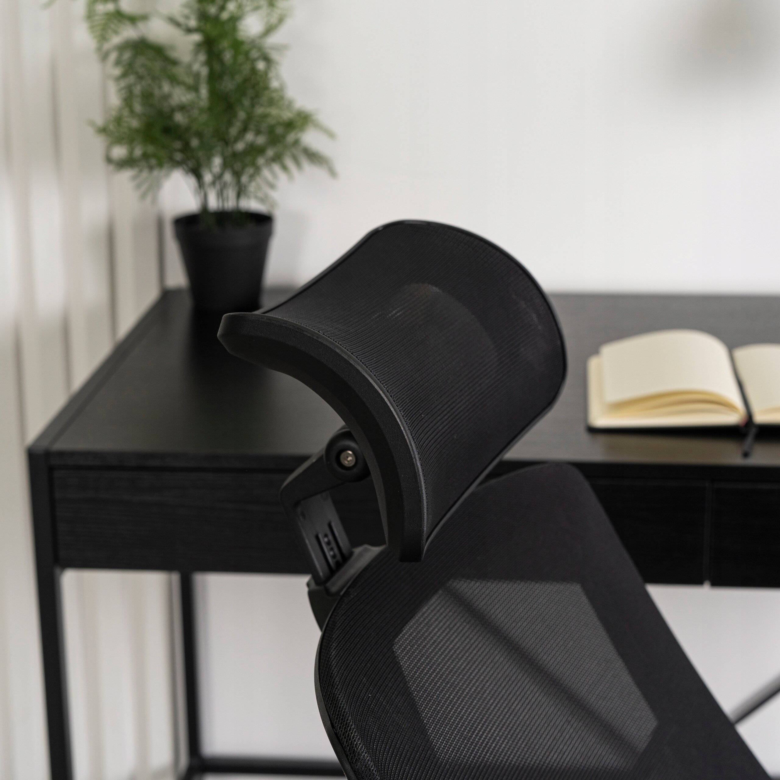 Czarny ergonomiczny fotel obrotowy z siatkowym oparciem, ustawiony przy czarnym biurku. Na biurku znajduje się otwarty notes i doniczka z zieloną rośliną.