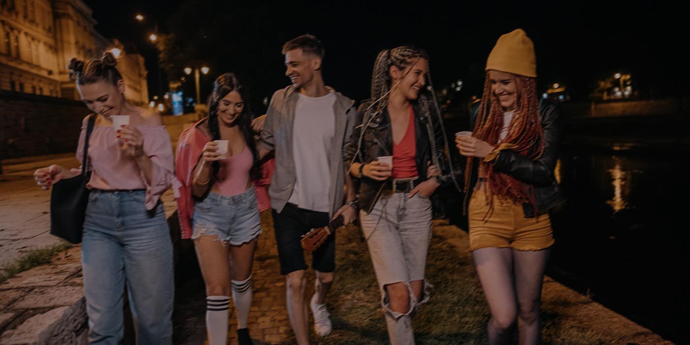 Pięcioro przyjaciół, cztery kobiety i jeden mężczyzna, spaceruje nocą wzdłuż rzeki, trzymając drinki pod latarniami. Pionowy suwak dzieli obraz, lewa strona jest przyciemniona i stonowana, a prawa jasna i kolorowa, odsłaniając wyraźniejsze detale, takie jak żółta czapka, podarte dżinsy i odbicia w wodzie.