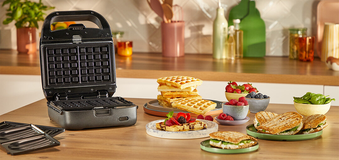 Opiekacz SW862B z płytami do gofrów, obok wymienne płyty i talerze z goframi, kanapkami i owocami.