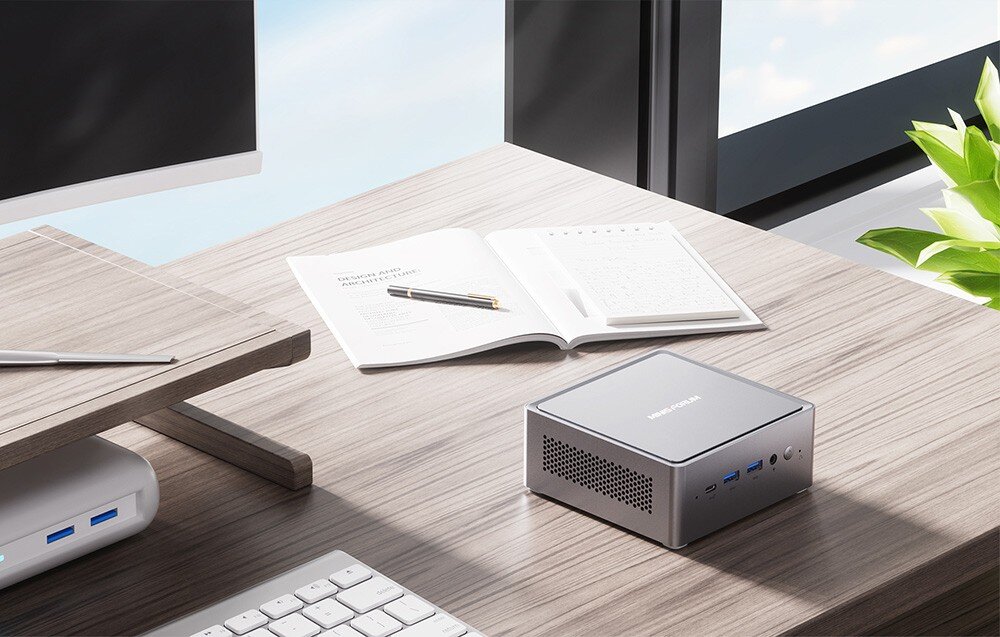 Komputer MINISFORUM NAB6 Lite Mini PC na drewnianym biurku przy oknie, obok otwartego notesu, klawiatury i monitora w jasnym biurze.