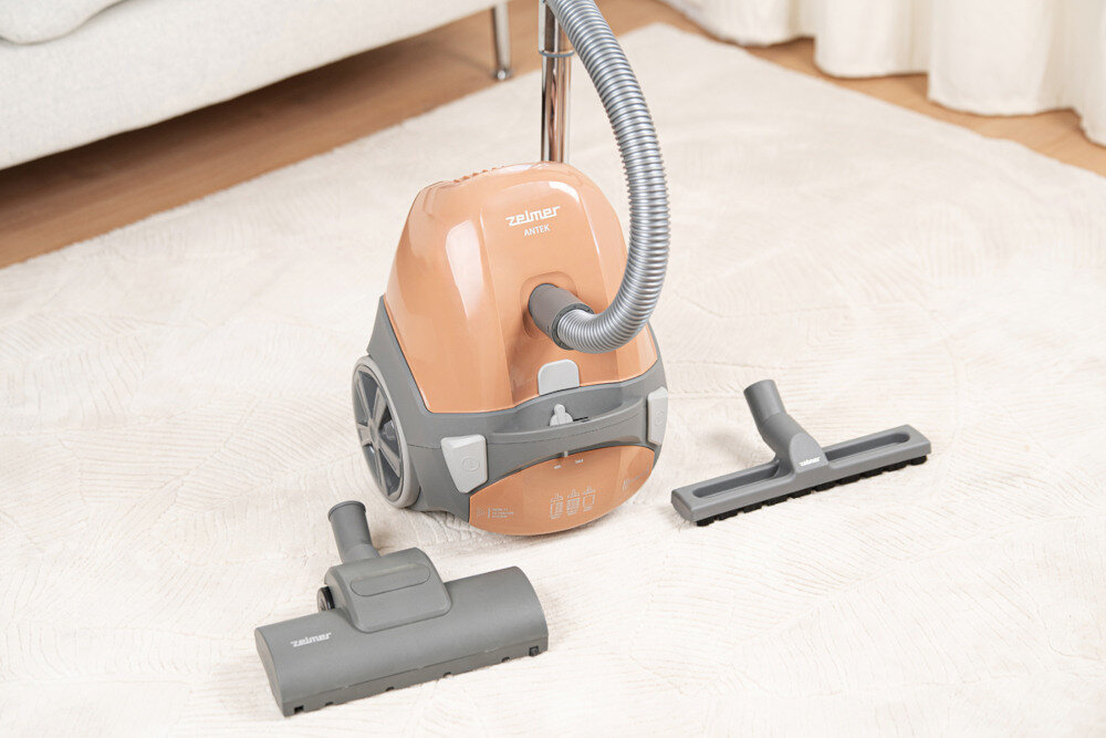 Na jasnym dywanie stoi odkurzacz w kolorze brązowym z szarymi elementami, z widocznym logo \'zelmer\' i napisem \'ANTEK\'. Obok odkurzacza leżą dwie szare końcówki ssące.