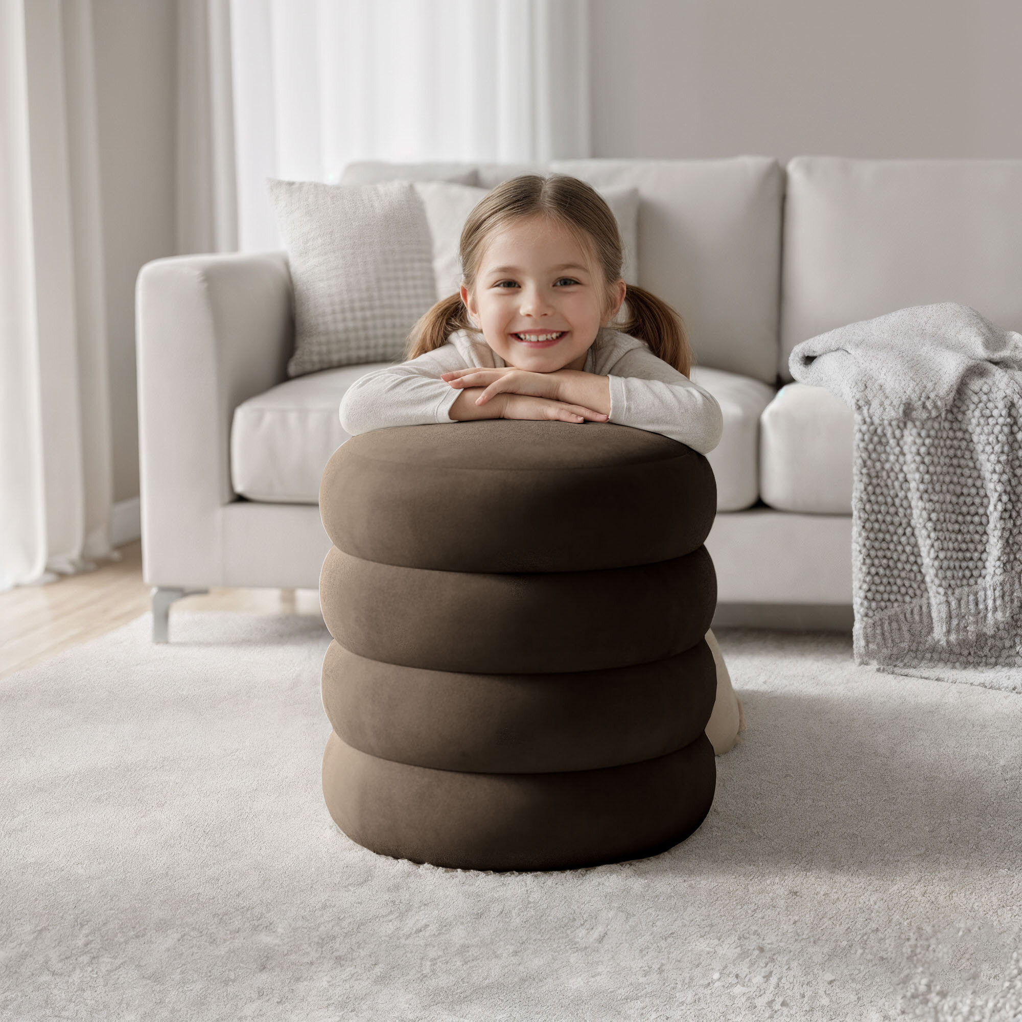 Na obrazie znajduje się brązowa, welurowa pufa o okrągłym kształcie, na której opiera się dziecko. W tle widoczna jest jasna sofa z poduszką i kocem.