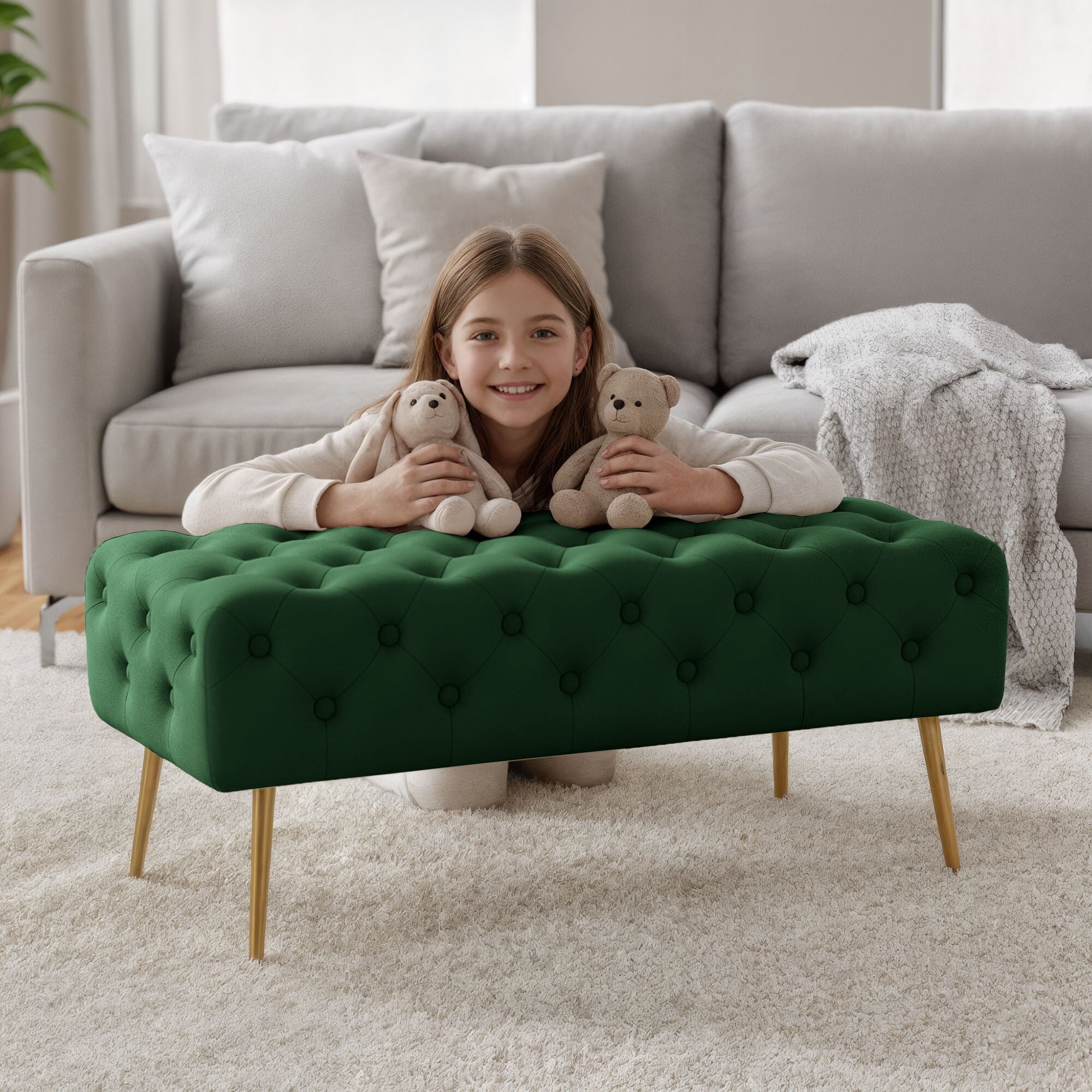 Na obrazie znajduje się tapicerowana ławka w kolorze butelkowej zieleni z pikowanym wzorem i złotymi nogami. W tle widoczna jest szara sofa i koc.