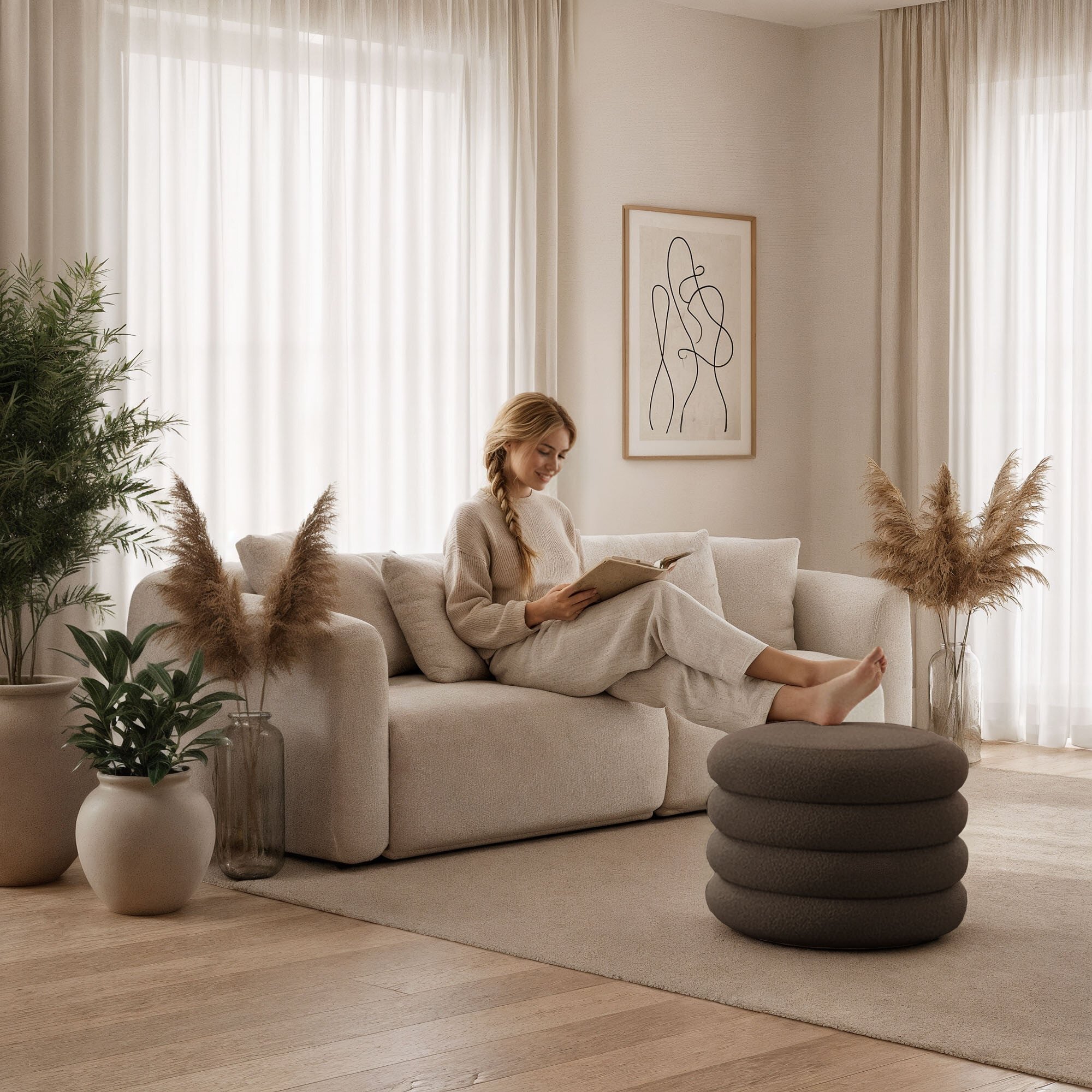 W jasnym, przytulnym wnętrzu znajduje się beżowa sofa, na której siedzi osoba czytająca książkę. Przed sofą stoi brązowa pufa o cylindrycznym kształcie, a w tle widać rośliny i abstrakcyjny obraz na ścianie.