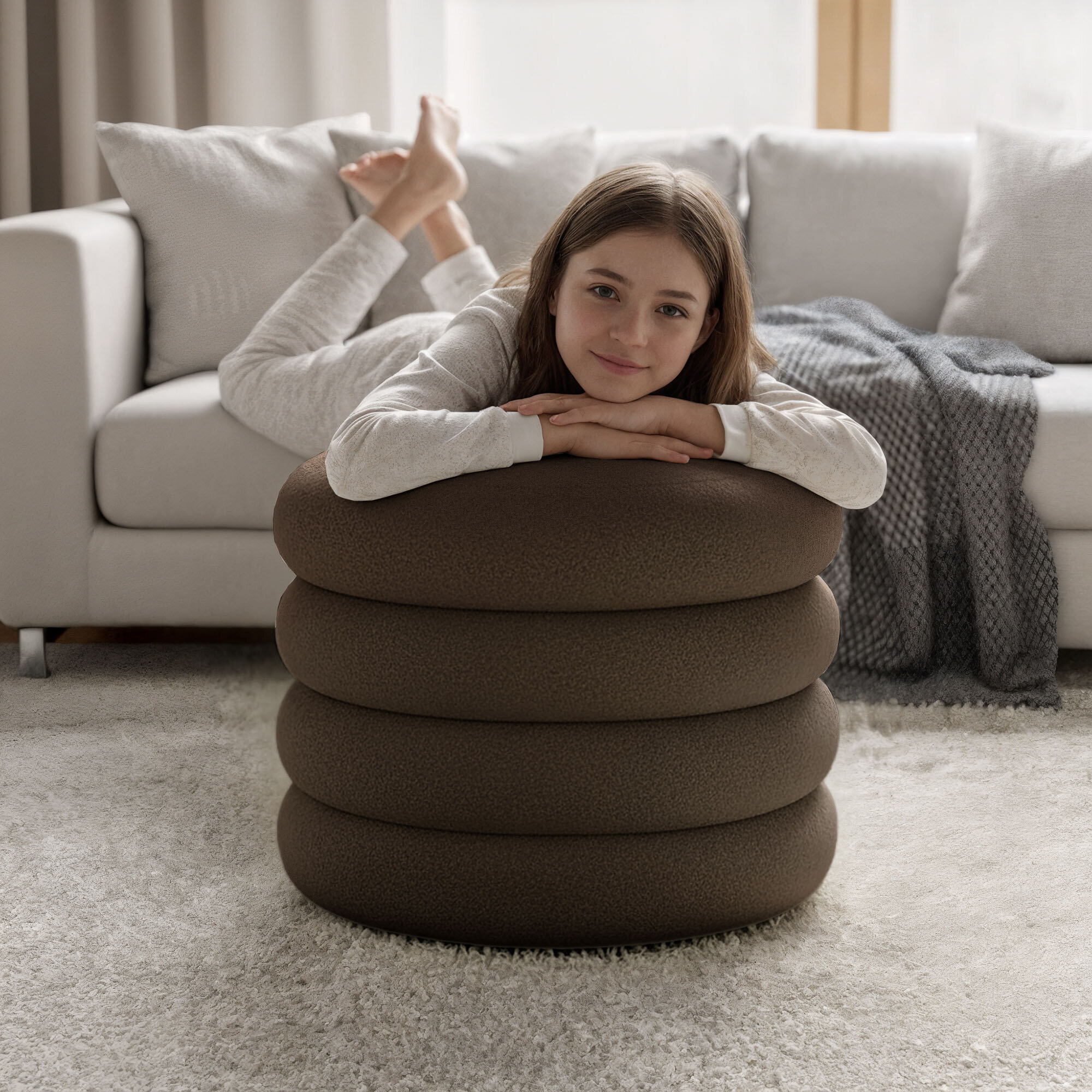 Brązowa pufa o okrągłym kształcie z segmentowaną strukturą, na której leży osoba. W tle znajduje się jasna sofa z poduszkami i kocem.