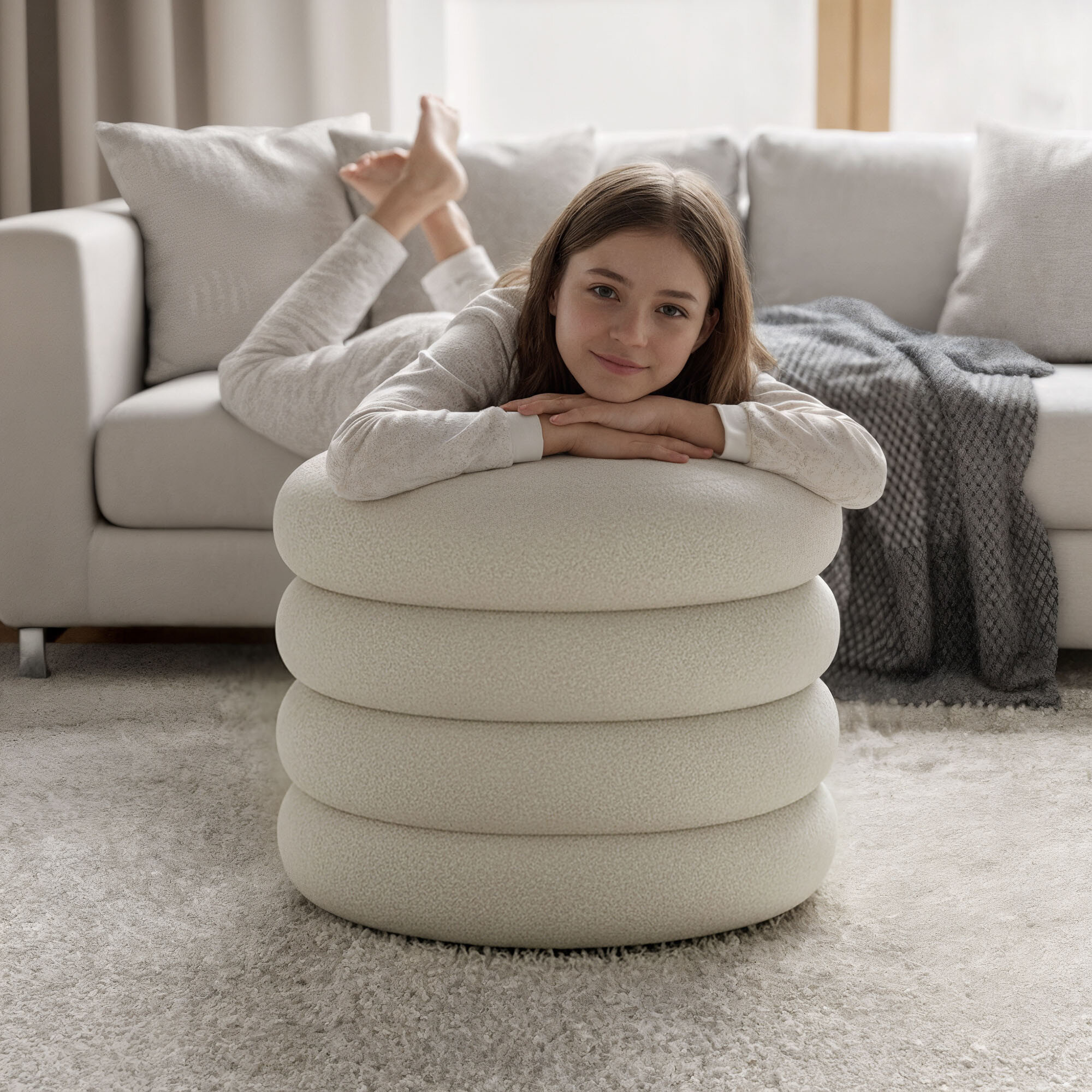 Dziewczynka leży na białej pufie o cylindrycznym kształcie, w tle znajduje się jasna sofa z poduszkami i kocem. Pomieszczenie jest jasne, z dużym oknem i dywanem na podłodze.